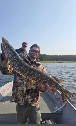 Man holding a large northern pike on a boat with another man standing behind him on a lake.