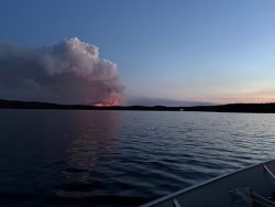 Smoke and flames from a wildfire rise behind a tree line at dusk, seen across a calm lake from a boat.