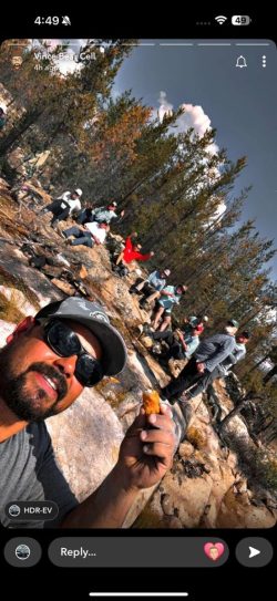 A group of people sit on rocks in a wooded outdoor area. One man in front holds food and takes a selfie.