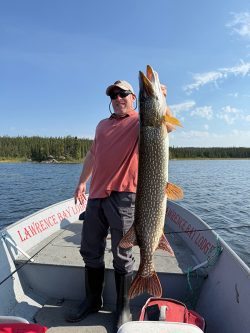 Man standing on a boat holding a large fish with both hands, lake and trees visible in the background.