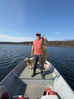 A man stands on a boat holding a large fish, with a dog beside him on a calm lake under a clear sky.