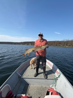 Man standing on a boat holding a large fish, with a dog beside him and a lake and forest in the background.