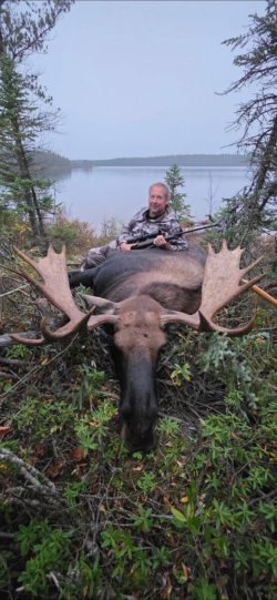 A person in camouflage sits behind a large moose lying on the ground in a forested area near a lake.