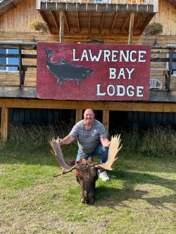 Man kneeling behind a moose antler trophy in front of a wooden building with a "Lawrence Bay Lodge" sign.
