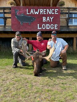 Three men pose with a moose in front of a "Lawrence Bay Lodge" sign outside a wooden building.