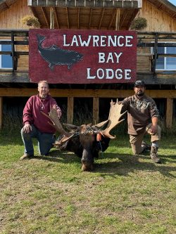 Two men kneel beside a moose head in front of a building with a "Lawrence Bay Lodge" sign.