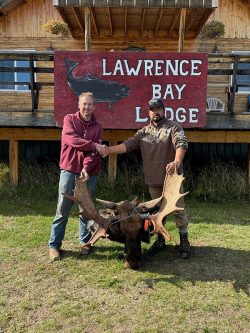 Two men shake hands in front of Lawrence Bay Lodge, standing behind a large moose antler trophy on the grass.