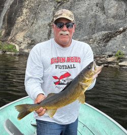Man in a boat holding a large fish, wearing sunglasses and a grey Eaton Reds Football shirt, with rocky background.