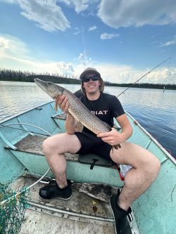 Person sitting in a boat holding a large fish, with fishing rod and net visible, on a lake under a partly cloudy sky.