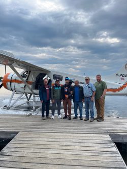 Six people stand on a dock in front of a floatplane on a cloudy day by the water.