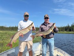 Two men standing in a boat on a lake, each holding a large fish with trees in the background.